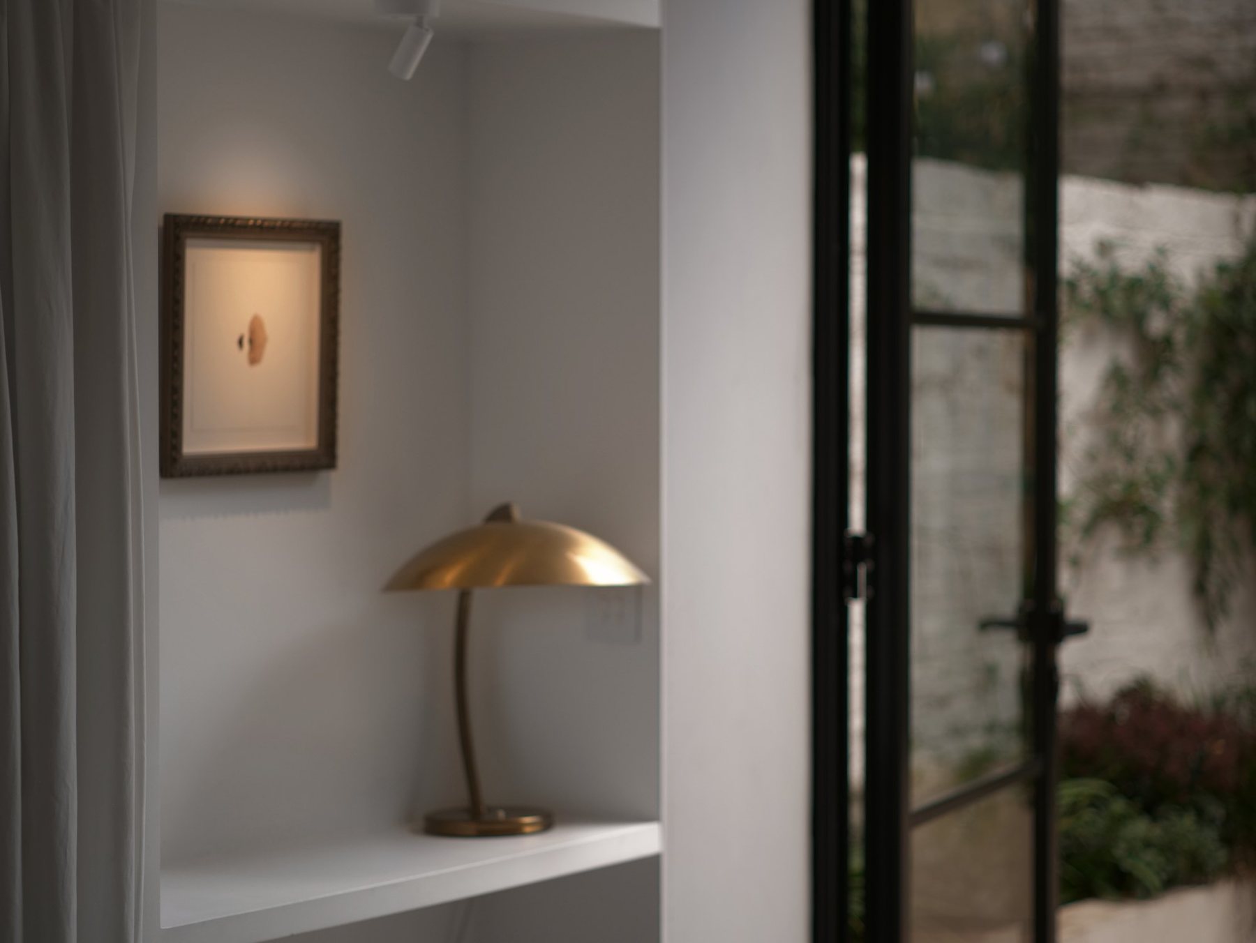 The hallway: a small framed work above a shelf and lamp, glass doors open to the garden.