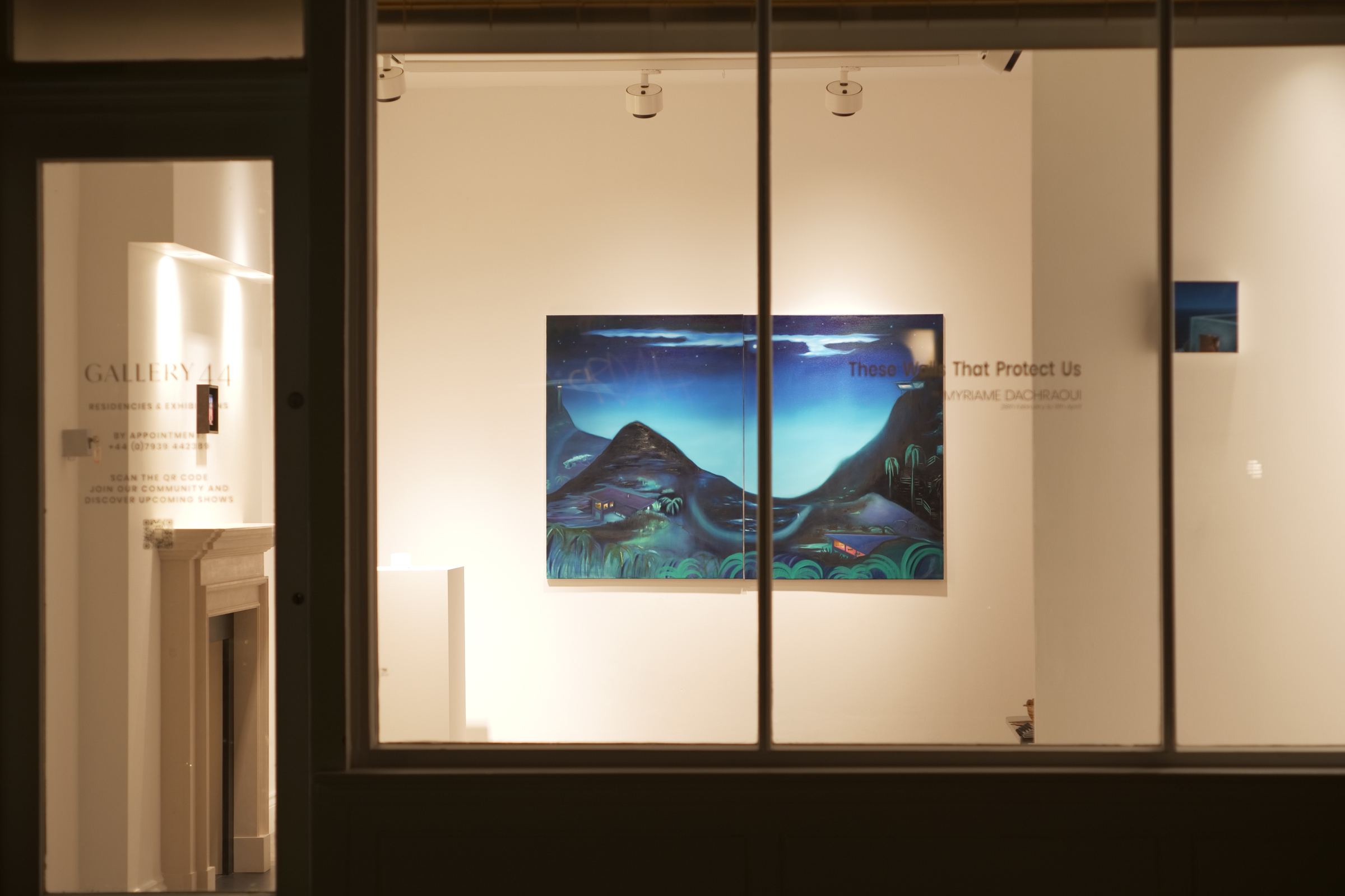 The illuminated gallery seen through the front window at night, with a blue diptych inside.