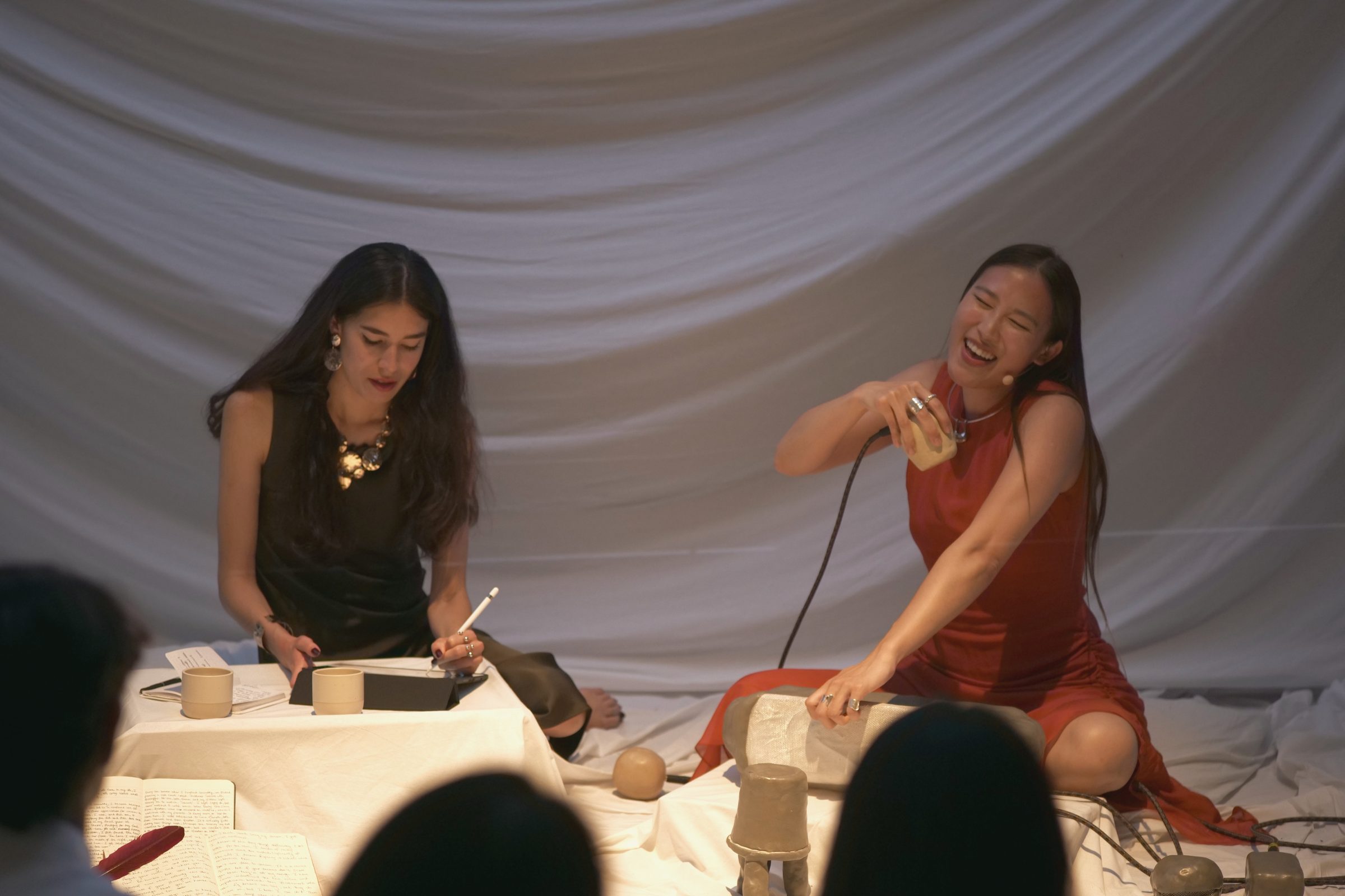 Hannah Dowling writes beside Betty C Fan, who plays a ceramic instrument, with an audience gathered around them.