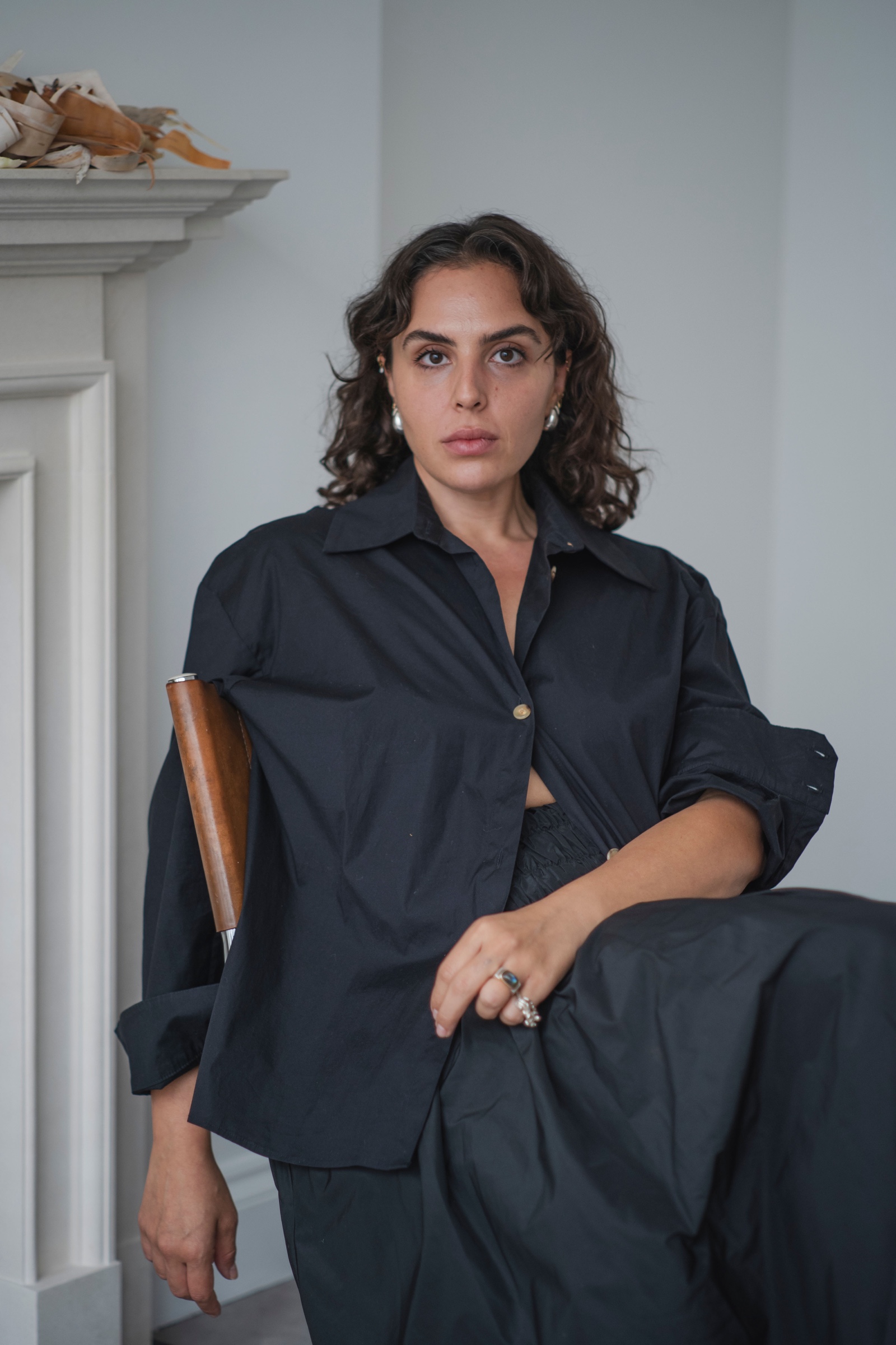Raya Kassisieh seated beside a white fireplace in a dark shirt and trousers, looking at the camera.