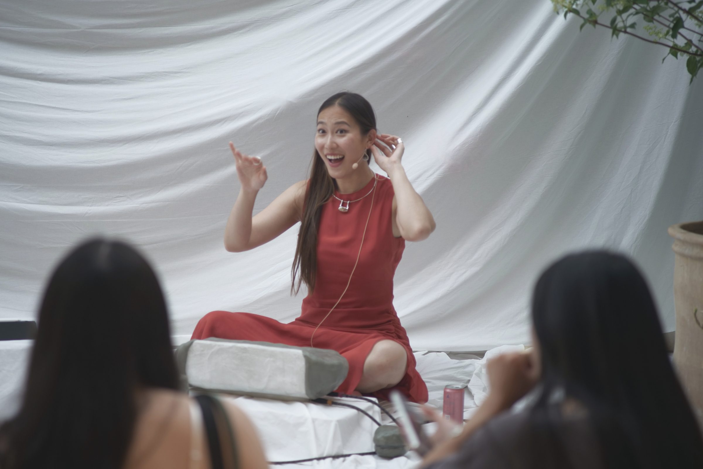 Betty C Fan, in a red dress, sits cross-legged on a white cloth, laughing and gesturing, the audience in the foreground.