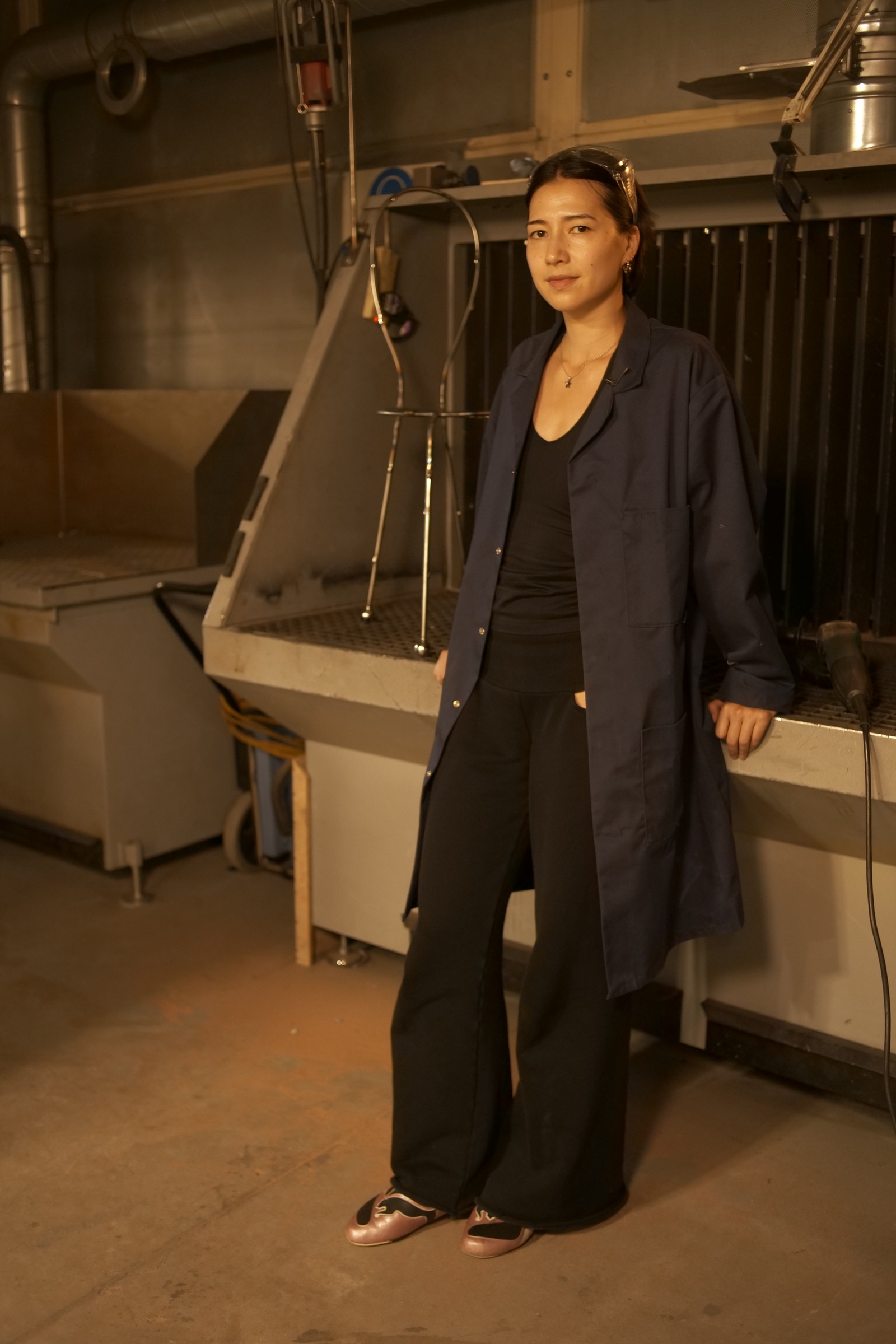 Marisa Müsing in a navy work coat and black top, with clear safety glasses pushed up on their head, standing in a metal workshop beside a steel chair frame and looking at the camera.