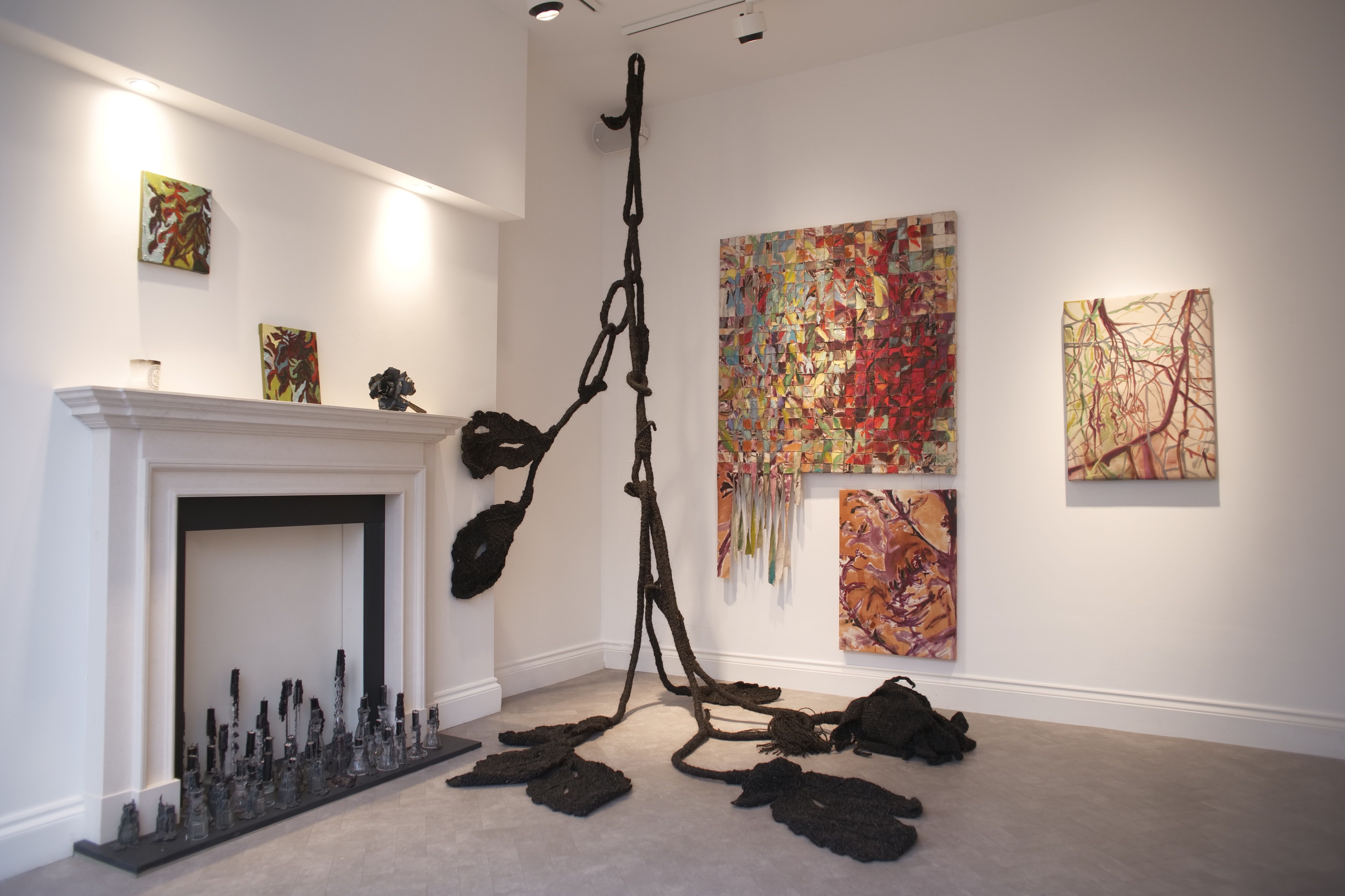 Installation view of Stasis: a suspended black textile sculpture beside a white fireplace, surrounded by woven canvases and paintings.