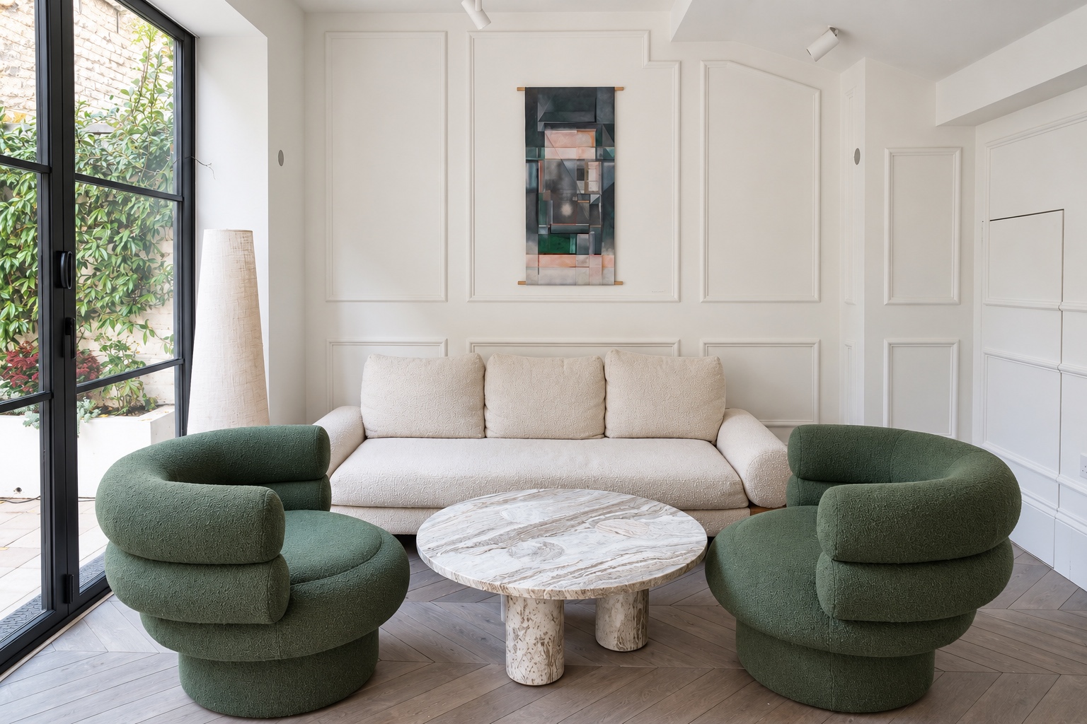The living room in daylight, with two green armchairs, a marble table, cream sofa, painting and garden doors.