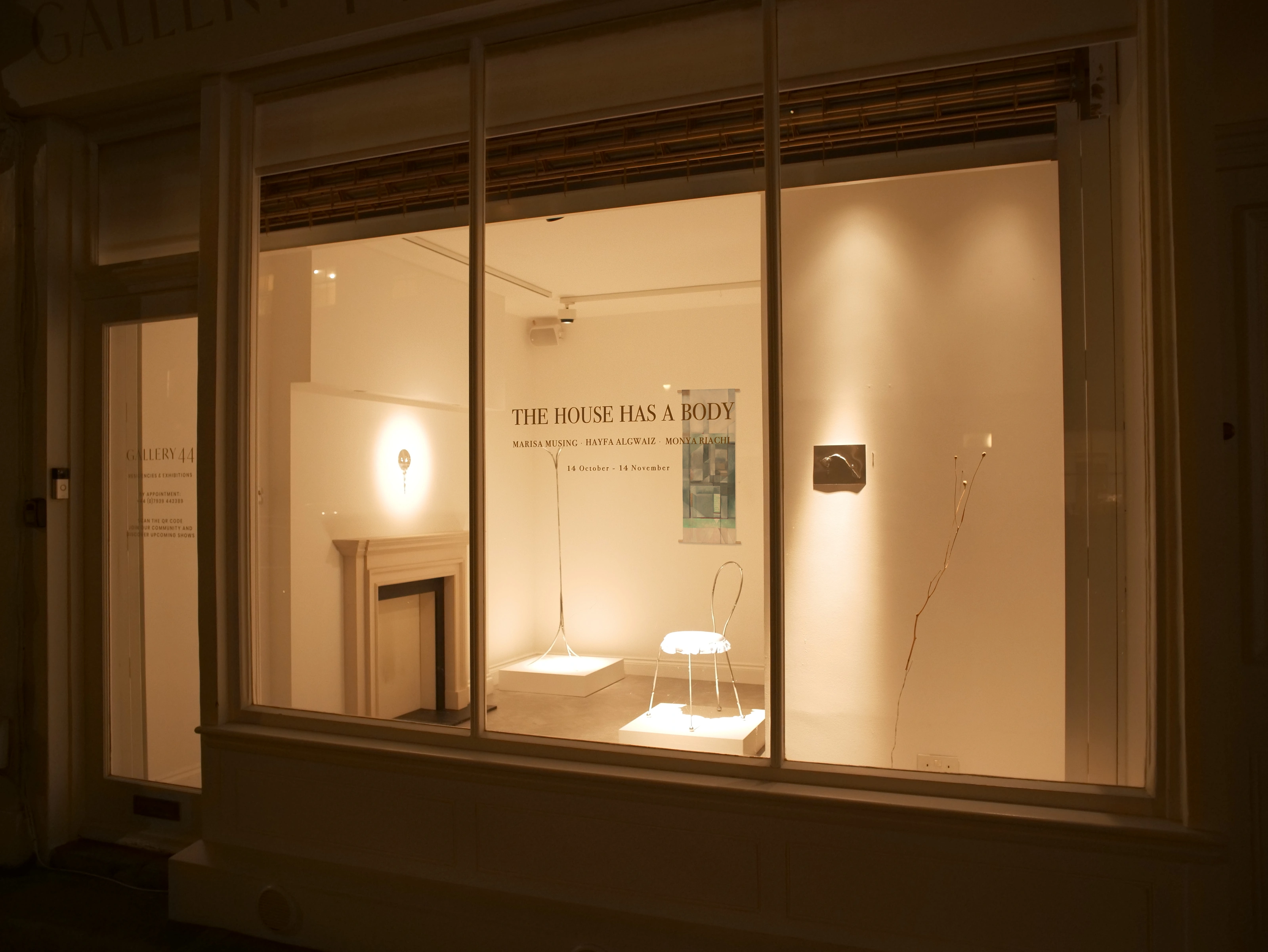 Gallery 44’s illuminated window at night, with The House Has a Body on the glass, Hayfa Algwaiz’s painting and sculptures inside.