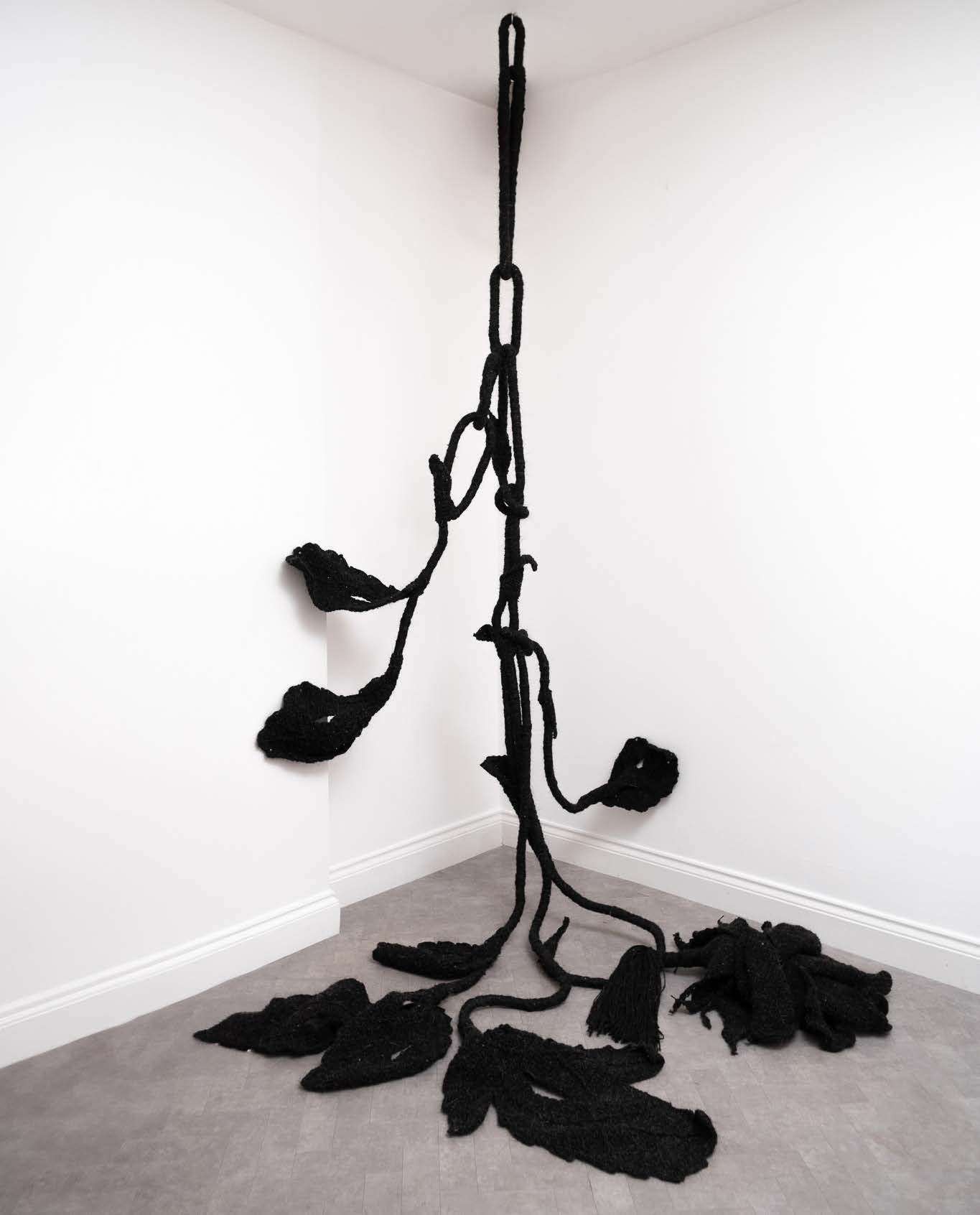 Black woollen roses and leaves on a long knotted stem, hanging from the ceiling into the corner of a white room and pooling on the floor.
