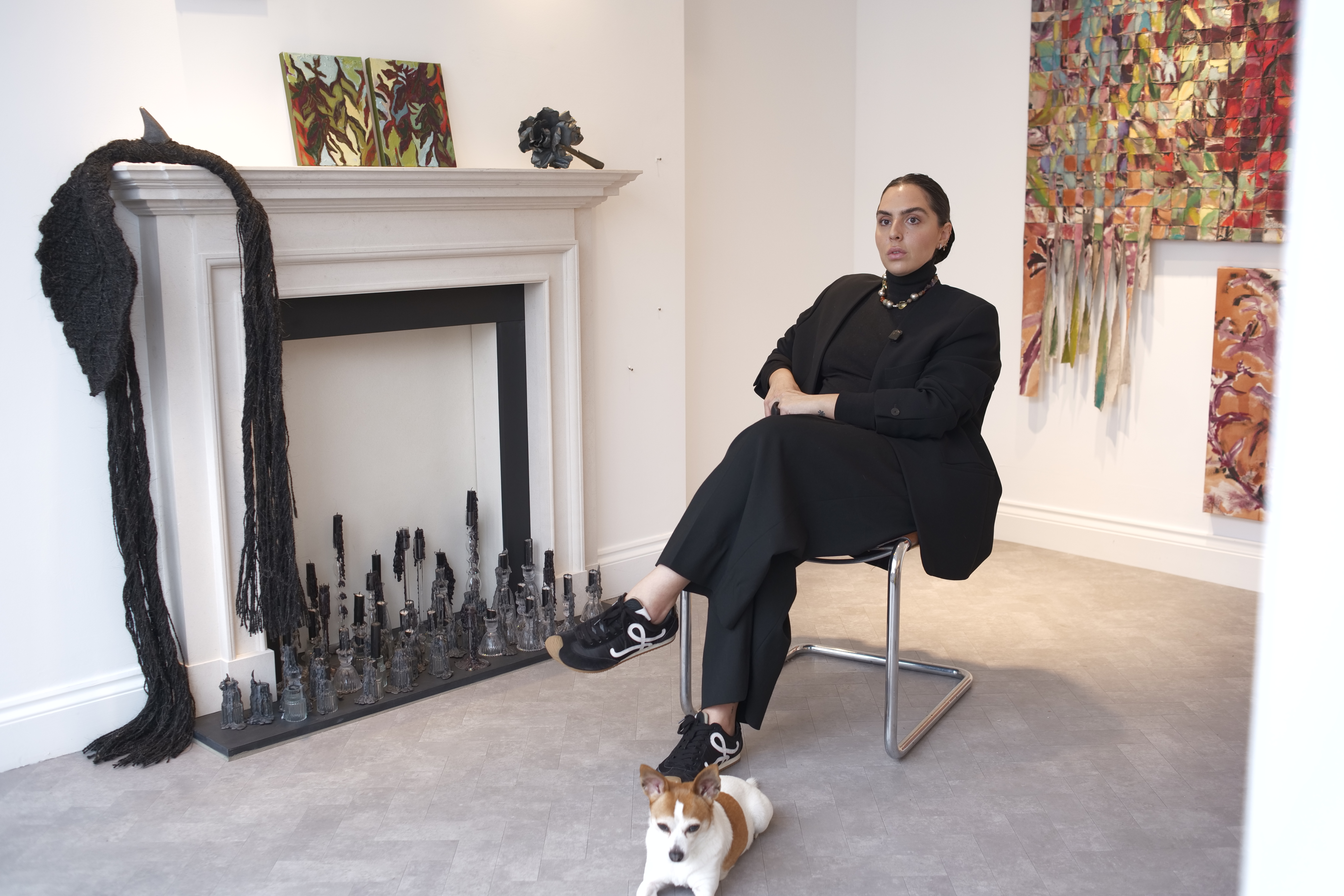 Raya Kassisieh seated beside the fireplace and her works in Stasis, with a small dog at her feet.