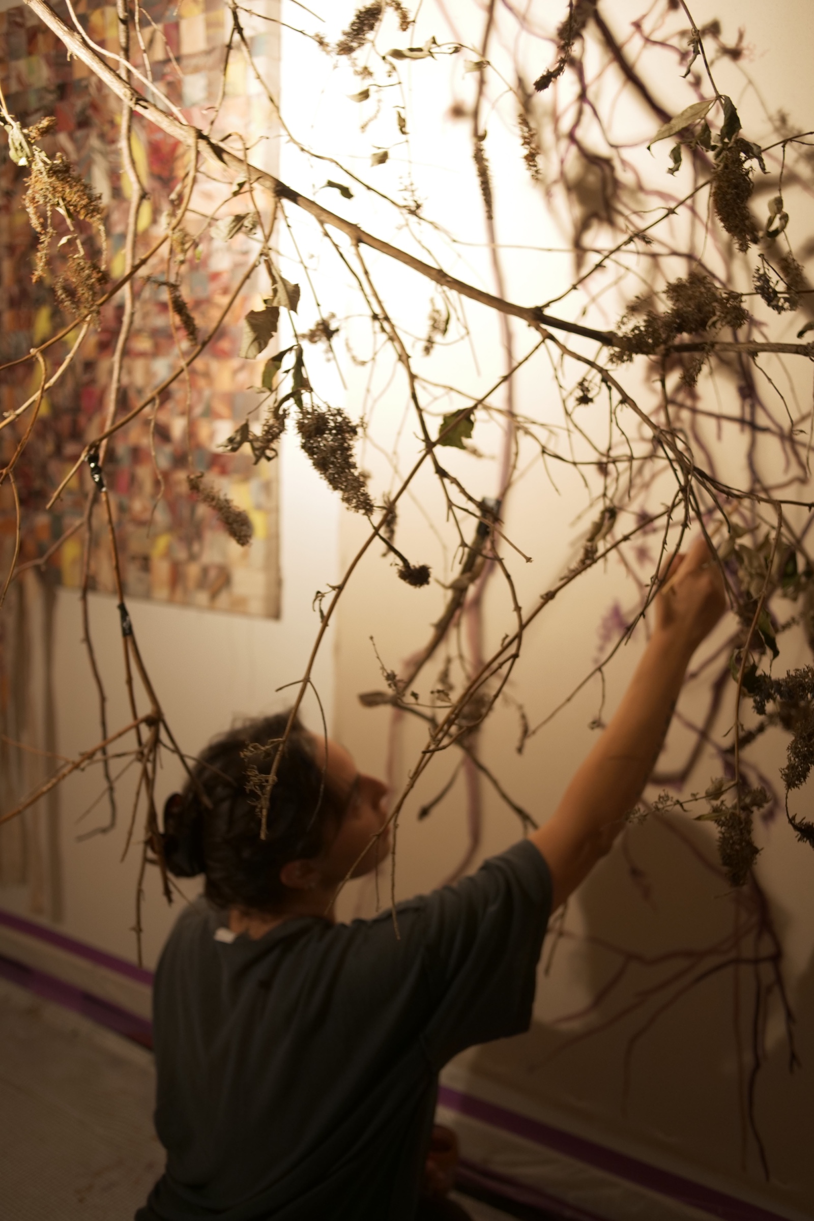 Raya Kassisieh reaches up through a suspended arrangement of branches while working in the studio, with woven canvases behind her.