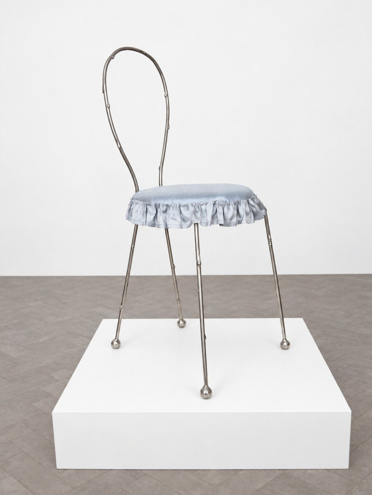 A slender steel chair with a looped back and a ruffled pale blue silk seat, on a white plinth.